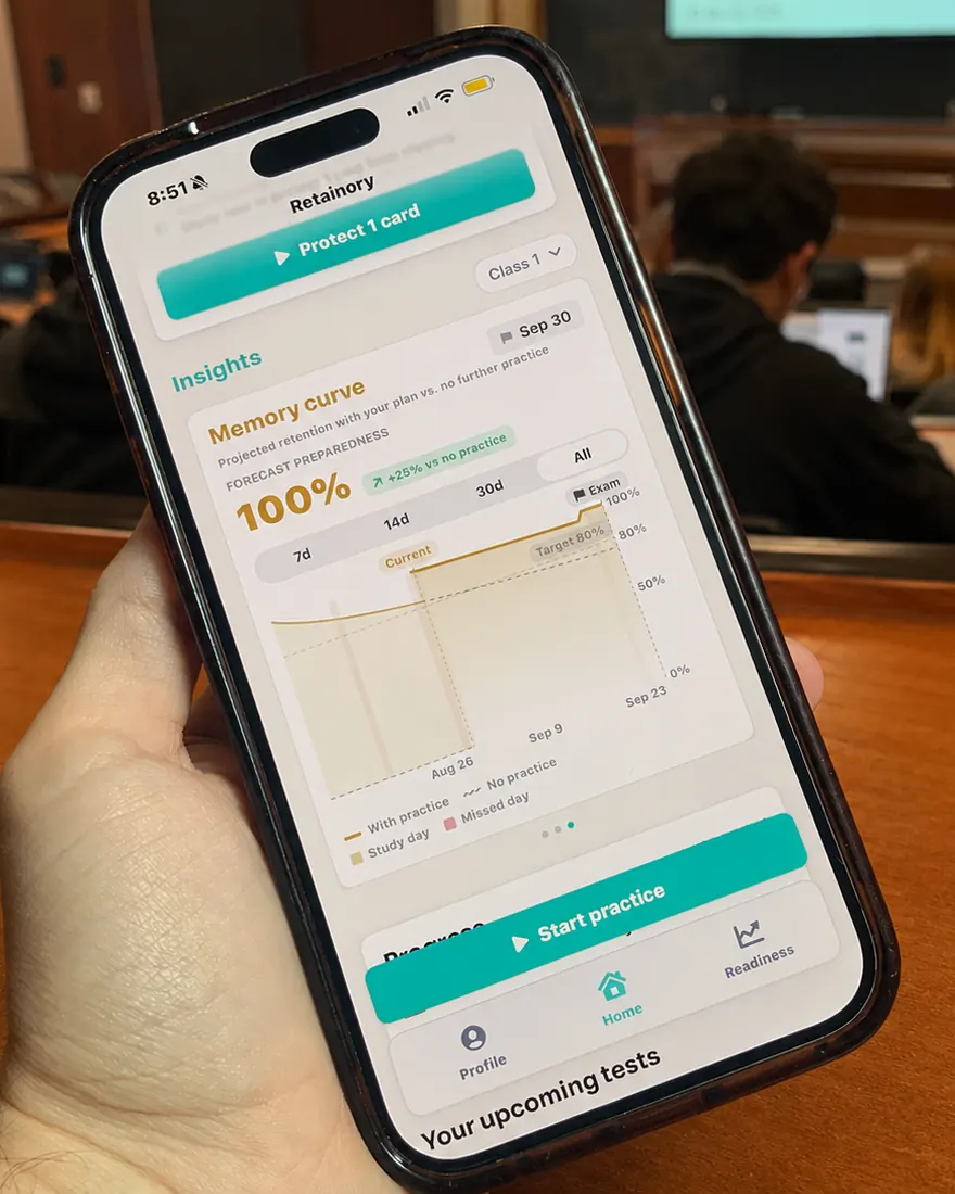 A hand holding a phone in a lecture hall, showing Retainory's insights view: a projected memory curve comparing retention with a study plan against no further practice, with the exam date marked on the timeline.
