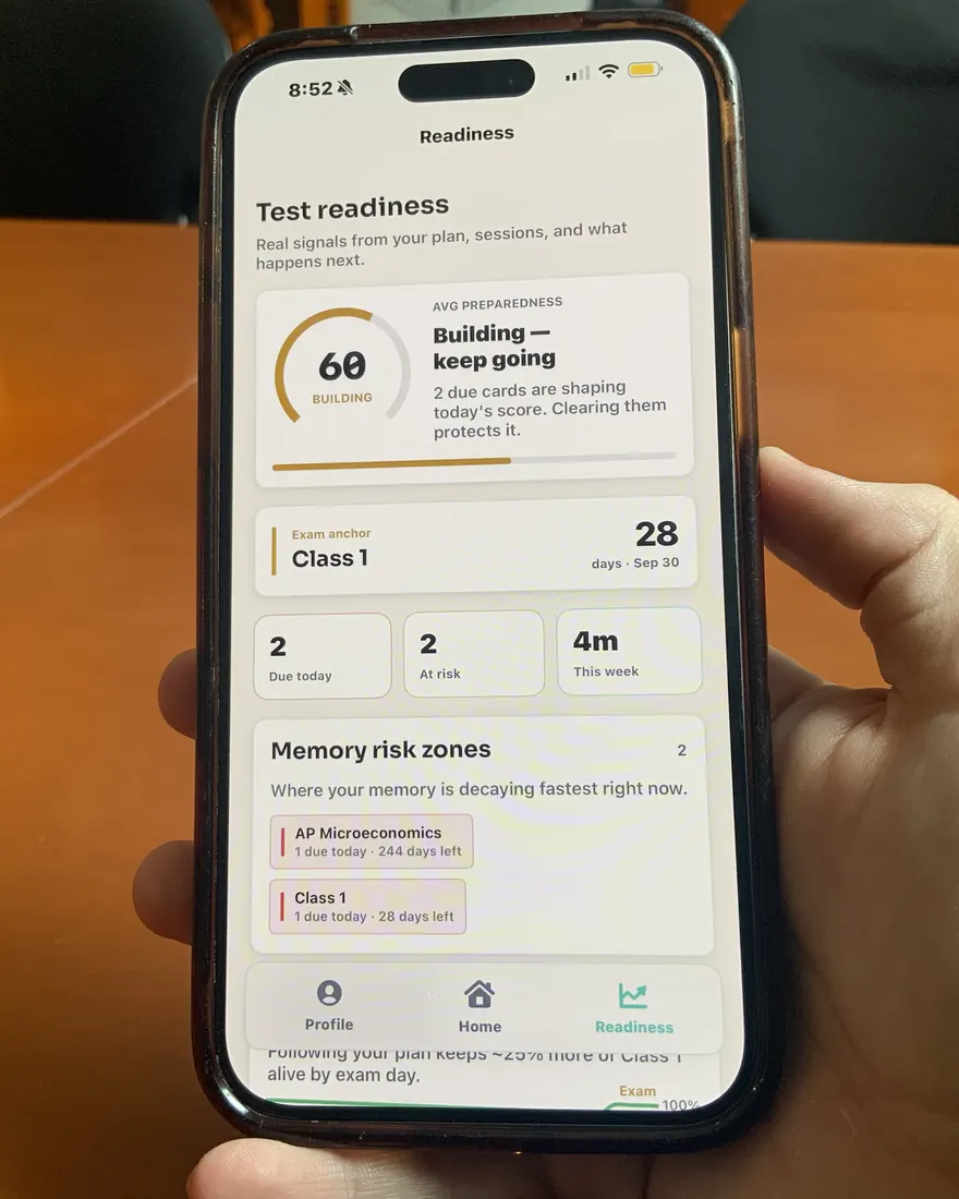 A hand holding a phone showing Retainory's readiness view: an average preparedness score of 60, labelled Building; an exam anchor of 28 days for Class 1; counts of 2 due today and 2 at risk; and a Memory risk zones list ranking AP Microeconomics and Class 1 by how fast each is decaying.